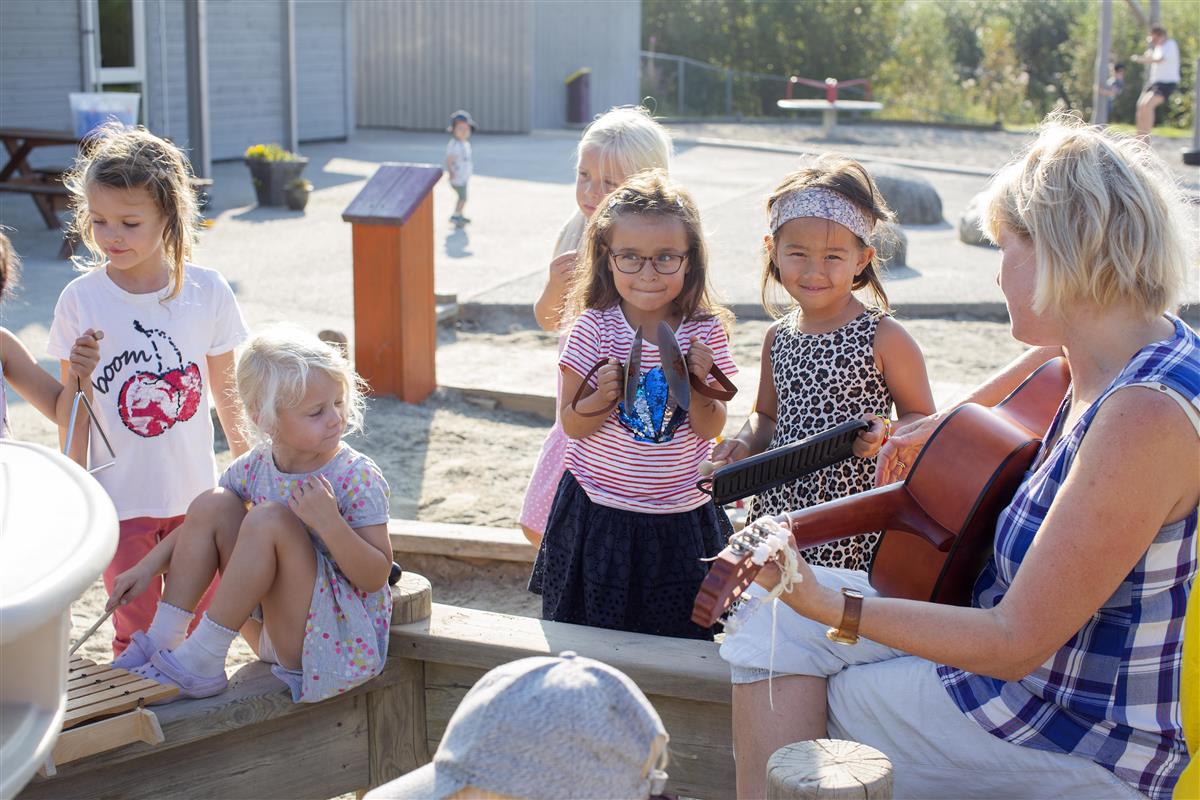 Foto av en barnehagelærer som spiller på gitar mens barna står rundt henne.  - Klikk for stort bilde