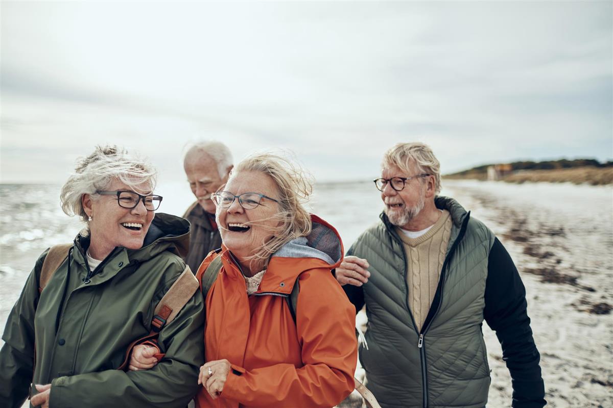 En gruppe eldre går tur på stranden, holder hverandre i armen og smiler. - Klikk for stort bilde